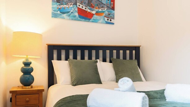 A neatly made bed with green bedding and white pillows, a bedside lamp, and a painting of boats on the wall. - Dingle Runda - 1 Bedroom (Dingle)