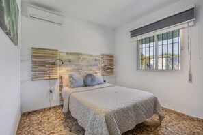 Um quarto com cama, janela com persianas, ar condicionado fixado na parede e piso texturizado.