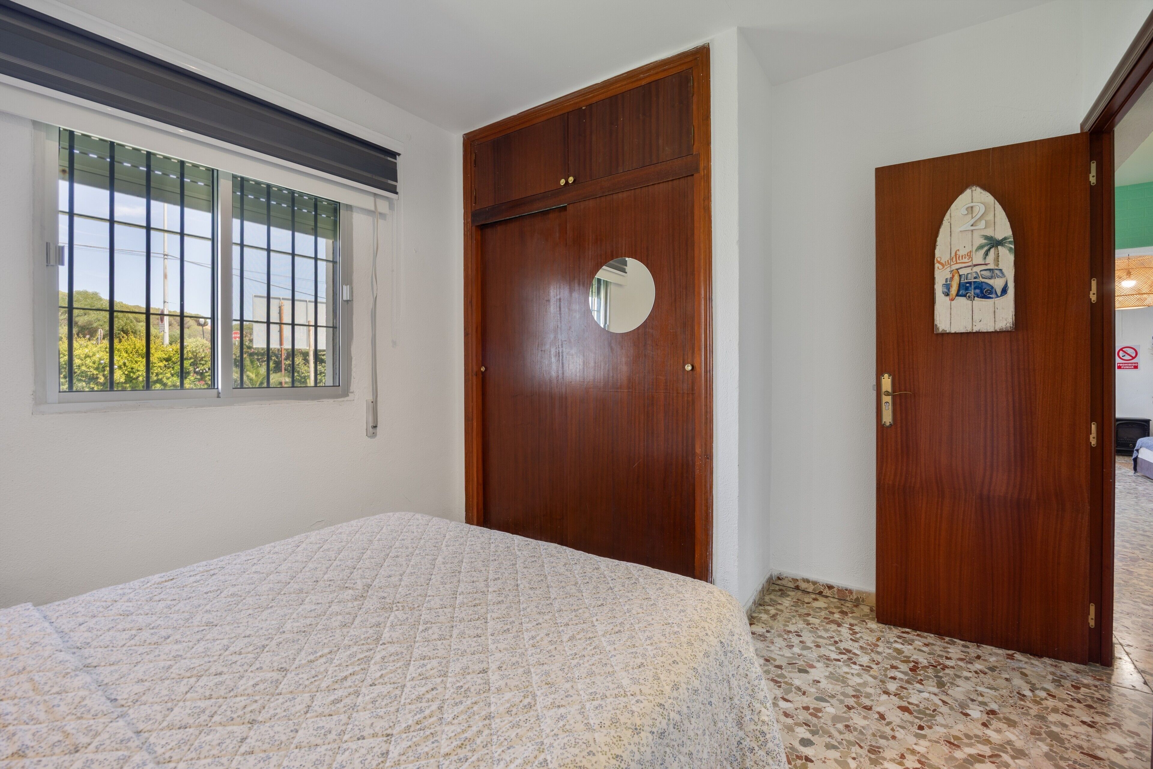 Un dormitorio con cama, ventana con persianas, puerta de madera con espejo y suelo de baldosas.