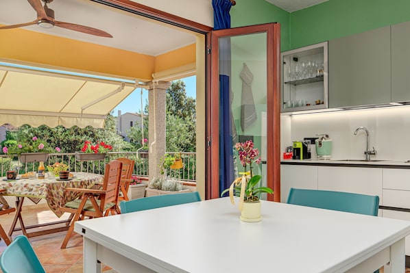 Un comedor con mesa y sillas, una cocina con lavamanos y armarios, y un balcón con plantas y vistas al exterior.