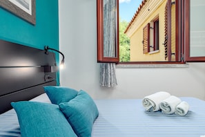 Una cama bien hecha con ropa de cama azul, una lámpara de noche y una ventana con contraventanas de madera.