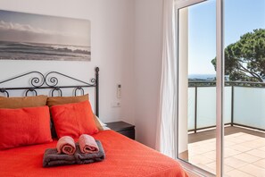 Un dormitorio con cama, almohadas, toallas, una mesita de noche, un enchufe en la pared y un balcón con vistas.