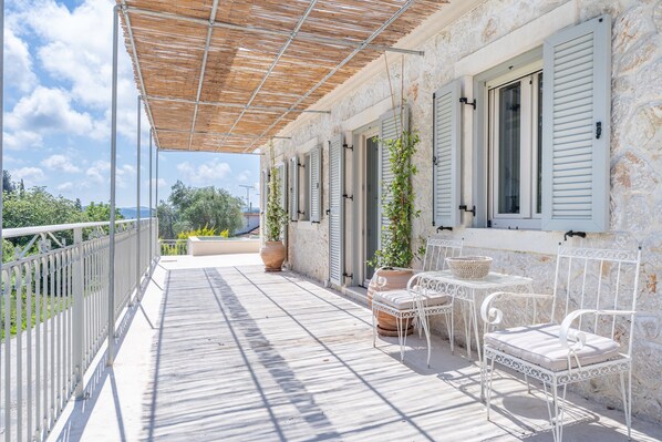Uma área externa coberta com guarda-corpo de metal branco, pérgola, plantas em vasos e cadeiras de metal branco.