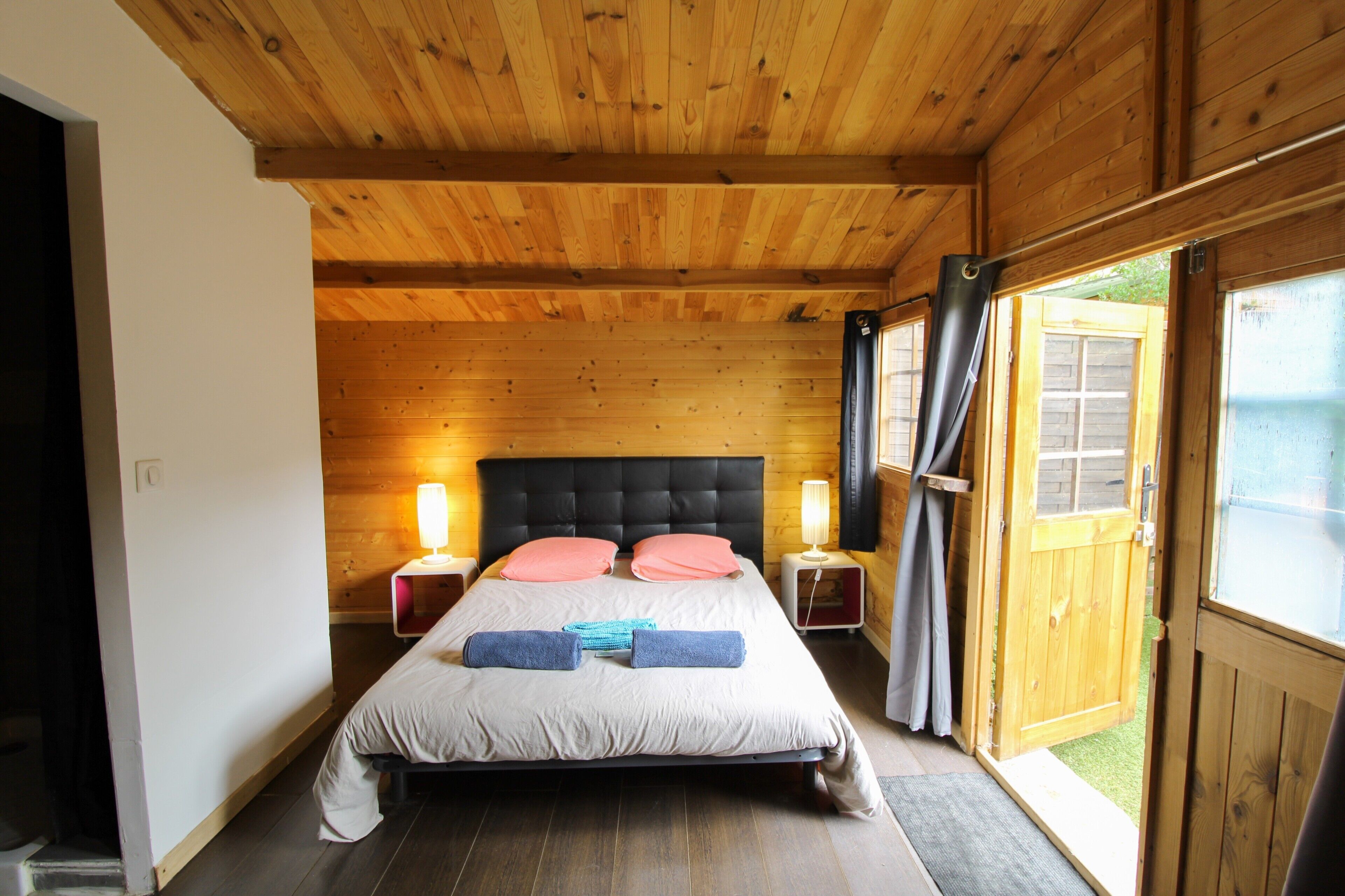 Un dormitorio con paneles de madera, una cama, dos lámparas de noche y vistas por una puerta abierta.