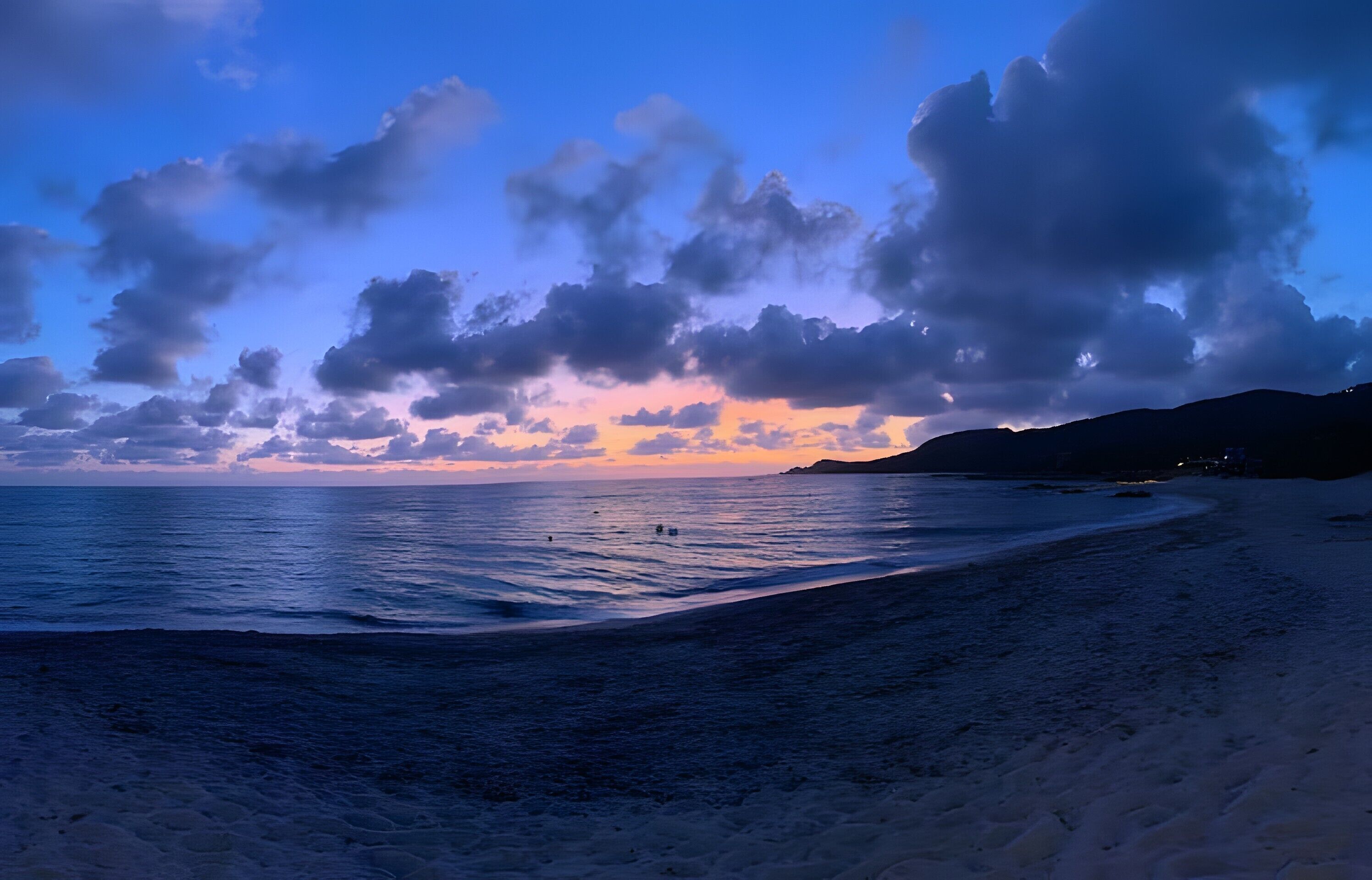 Uma praia com pôr do sol, águas calmas e colinas ao longe.