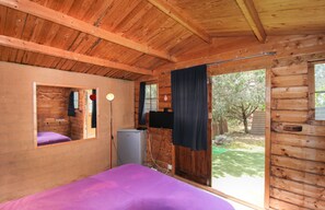 A log cabin room with a bed, a small refrigerator, a television, and a window with curtains.