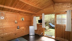Une cabane en bois comprenant un lit, un petit réfrigérateur, une télévision et une porte-fenêtre coulissante donnant sur un jardin.