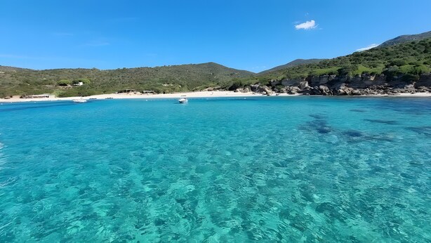 Une mer d’un bleu turquoise limpide, avec un bateau, une plage de sable et des collines verdoyantes en arrière-plan.