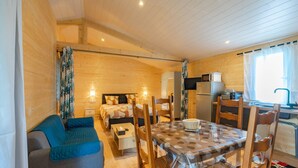 Ein gemütliches Blockhaus mit Holzvertäfelung, einem Essbereich mit einem gemusterten Tisch, einer Küchenzeile und einem Schlafzimmer mit Bett und Nachttisch.