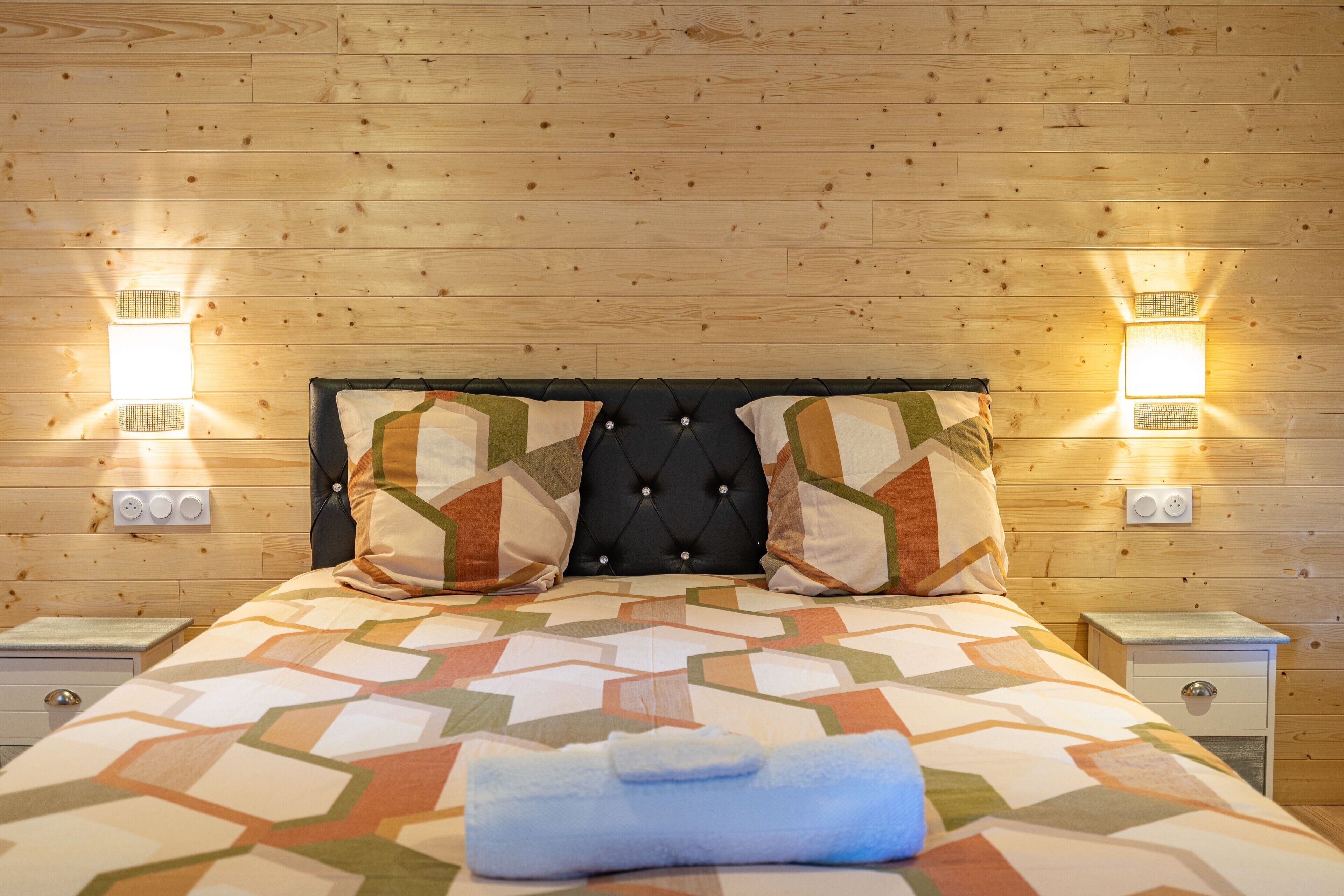A wooden-paneled room with a bed, two wall-mounted lights, and a nightstand with a drawer.