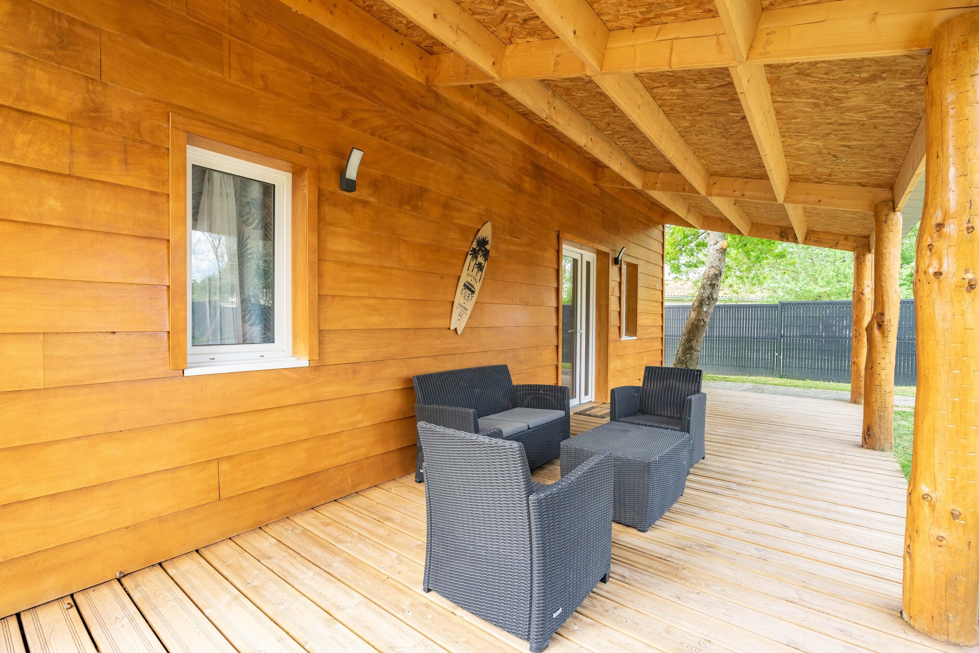 Een overdekt houten terras met rieten meubilair en een surfplank.