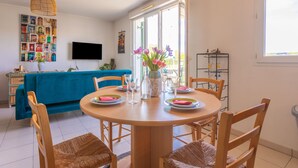 A dining area with a round table, chairs, a vase with flowers, a television on the wall, and a blue sofa in the background.