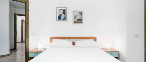 A bedroom with a bed, bedside tables, a wooden headboard, two framed artworks on the wall, and a door leading to another room.