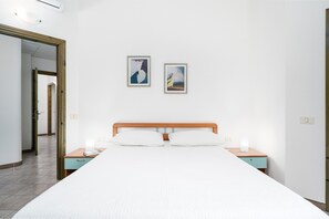 A bedroom with a bed, bedside tables, a wooden headboard, two framed artworks on the wall, and a door leading to another room.