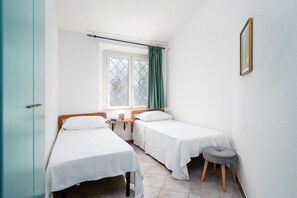 A room with two single beds, a window with curtains, a small stool, and a framed picture on the wall.