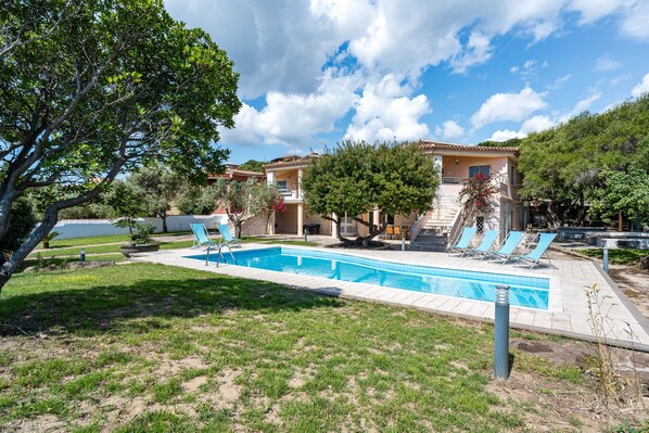 A swimming pool with lounge chairs, a building with a balcony, and surrounding greenery.