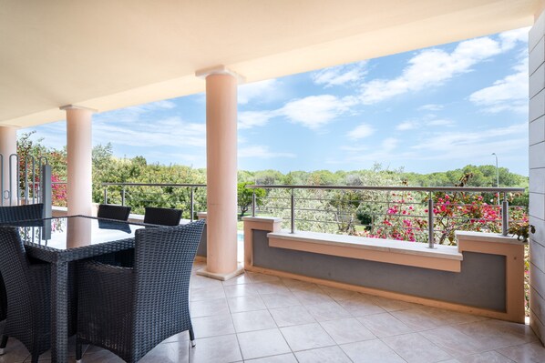 A covered outdoor area with a table and chairs, a railing, and a view of greenery.