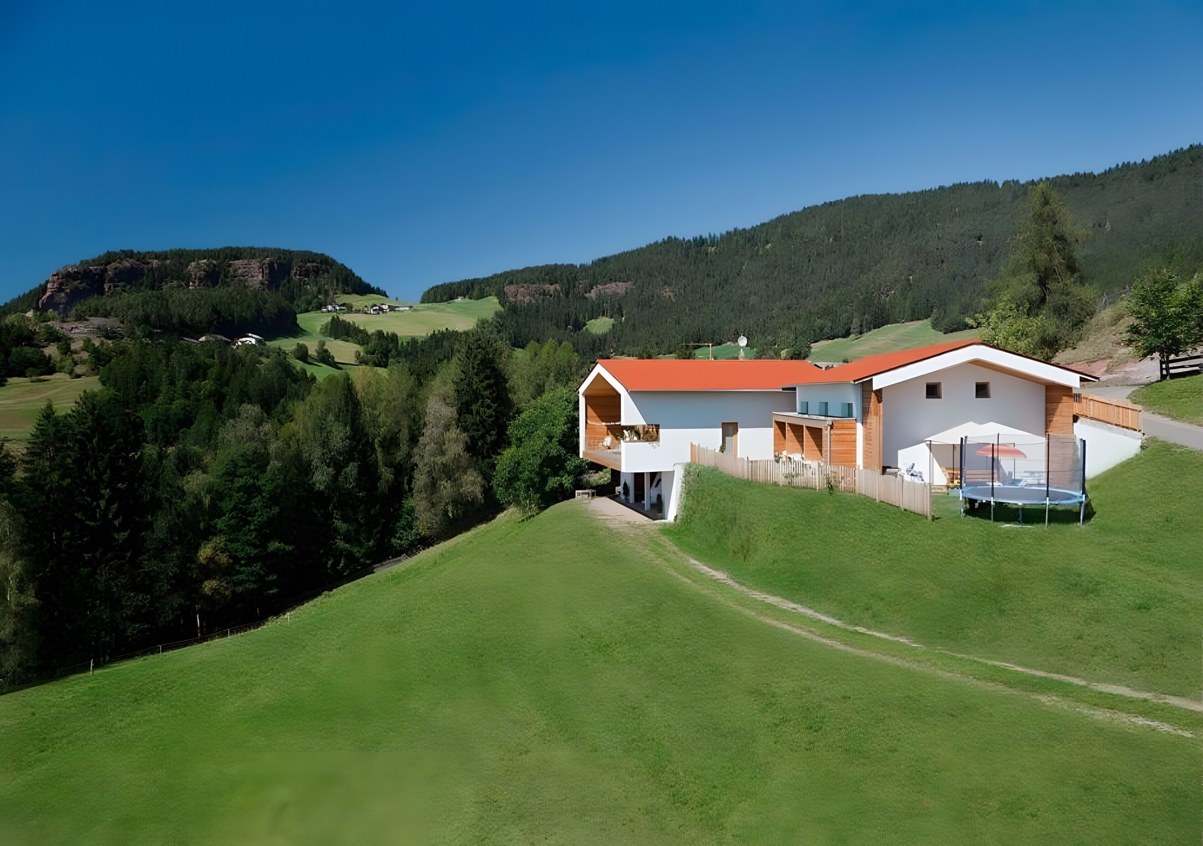 Une maison moderne avec un toit rouge, un balcon et un trampoline dans le jardin, entourée de collines verdoyantes et d’arbres.