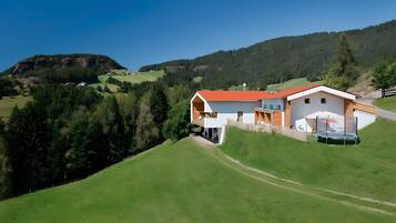 Une maison moderne avec un toit rouge, un balcon et un trampoline dans le jardin, entourée de collines verdoyantes et d’arbres.