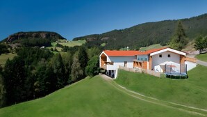 A modern house with a red roof, a balcony, and a trampoline in the backyard, surrounded by green hills and trees.