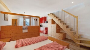 A bedroom with a wooden headboard, a bed with checkered bedding, a wooden staircase, and a kitchen area with red cabinets.