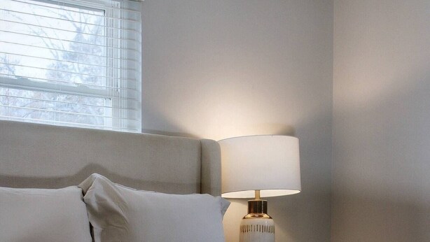 A neatly made bed with a headboard, a bedside lamp, and a window with blinds. - Charming 2-bedroom townhome in Raleigh with AC, WiFi (Raleigh)