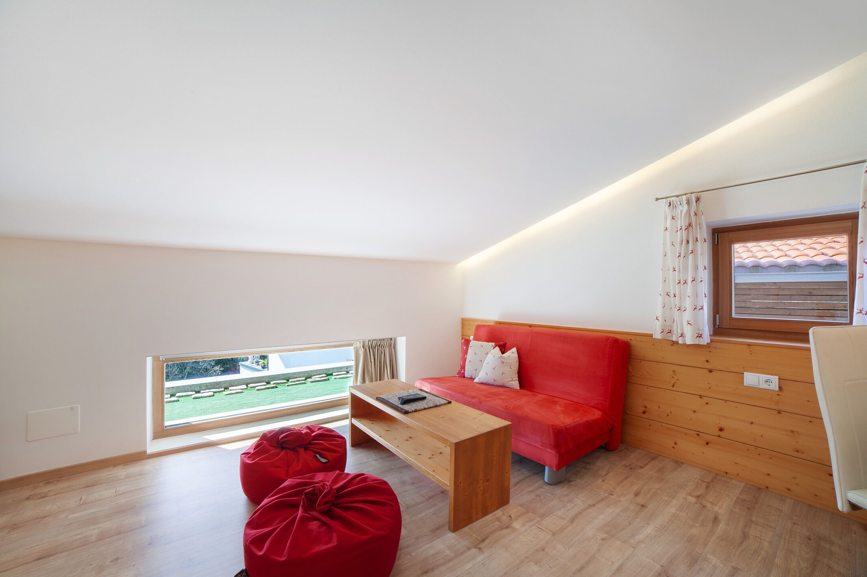 A modern room with a wooden floor, a red sofa, a wooden coffee table, a red bean bag, and a window with a view of a green landscape.