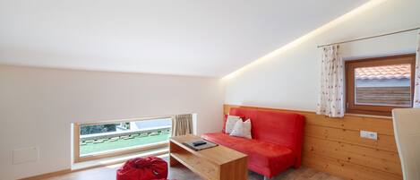 A modern room with a wooden floor, a red sofa, a wooden coffee table, a red bean bag, and a window with a view of a green landscape.