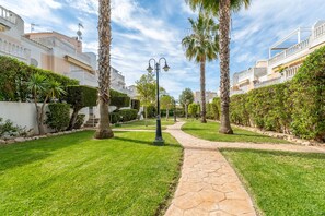 A pathway leading through a well-maintained garden with palm trees and neatly trimmed hedges, flanked by buildings with balconies and satellite dishes. - Apartment 'A Un Paso Del Mar' with Shared Pool and Balcony (Guardamar del segura)
