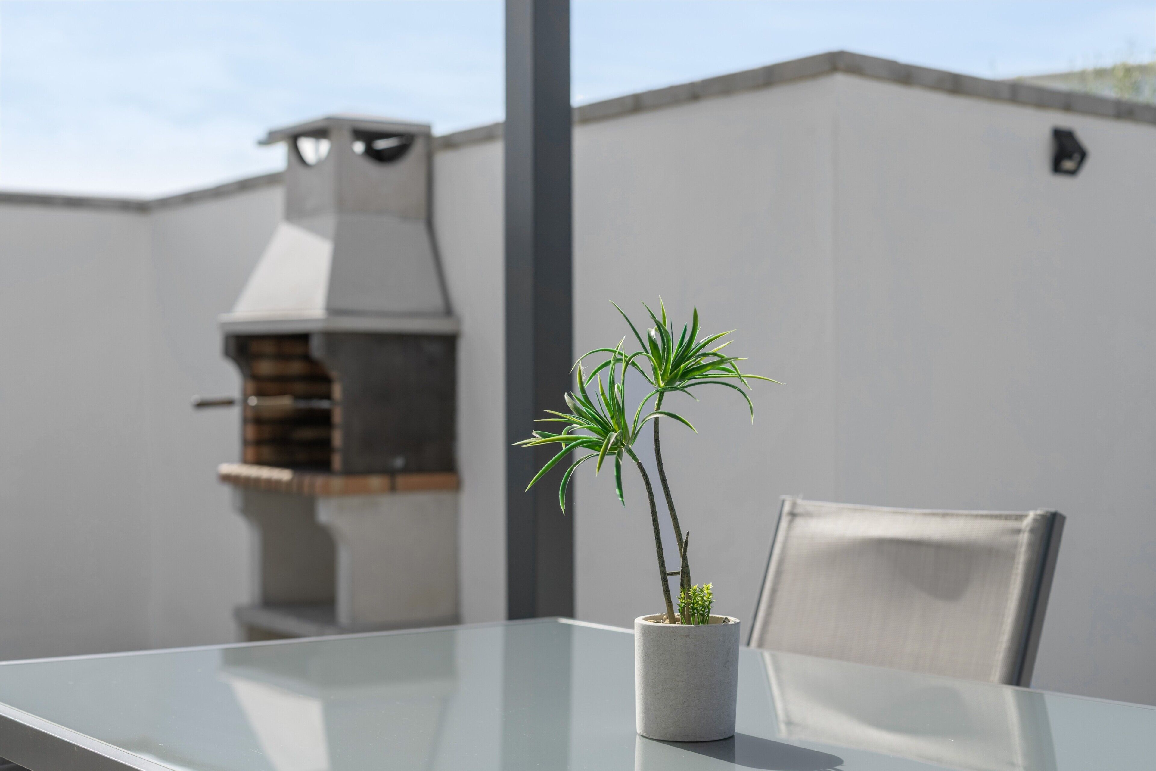 Et moderne udendørs opholdsområde med et glasbord, en potteplante og en grill i baggrunden.