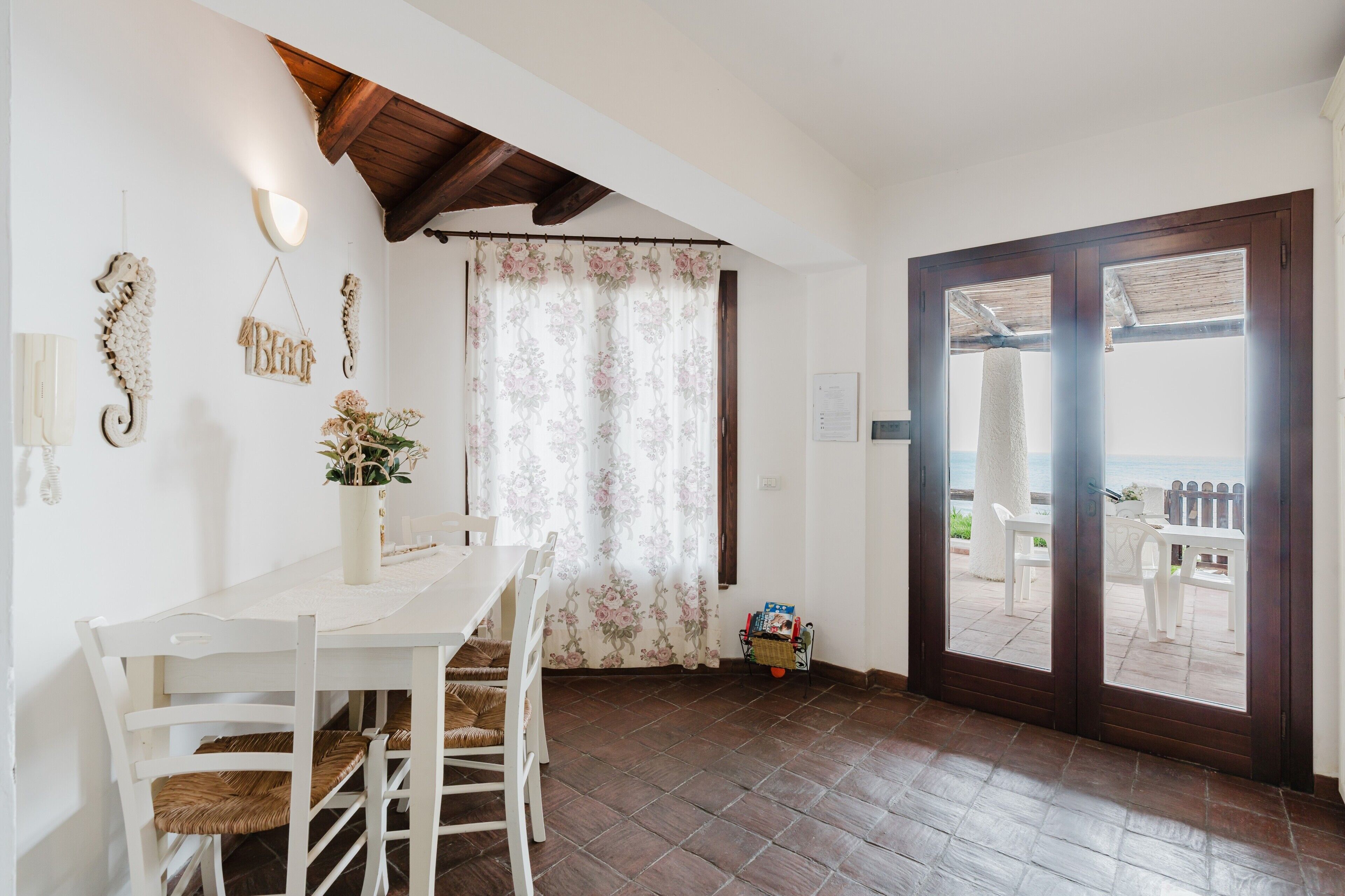 A dining area with a white table and chairs, a wooden ceiling, a floral curtain, and a view through a door to an outdoor patio.