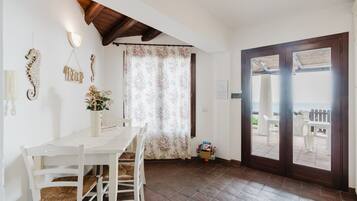 A dining area with a white table and chairs, a wooden ceiling, a floral curtain, and a view through a door to an outdoor patio.