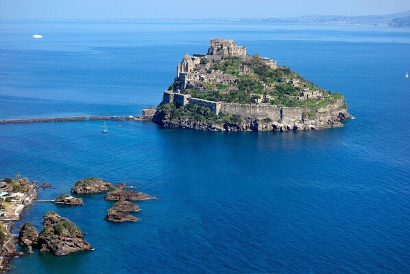 A coastal castle surrounded by clear blue waters, rocky outcrops, and a distant cityscape.