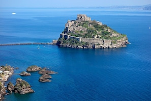 A coastal castle surrounded by clear blue waters, rocky outcrops, and a distant cityscape.