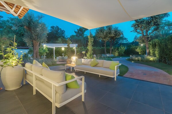 An outdoor patio with a white sofa, a large potted plant, and a pergola with lights.