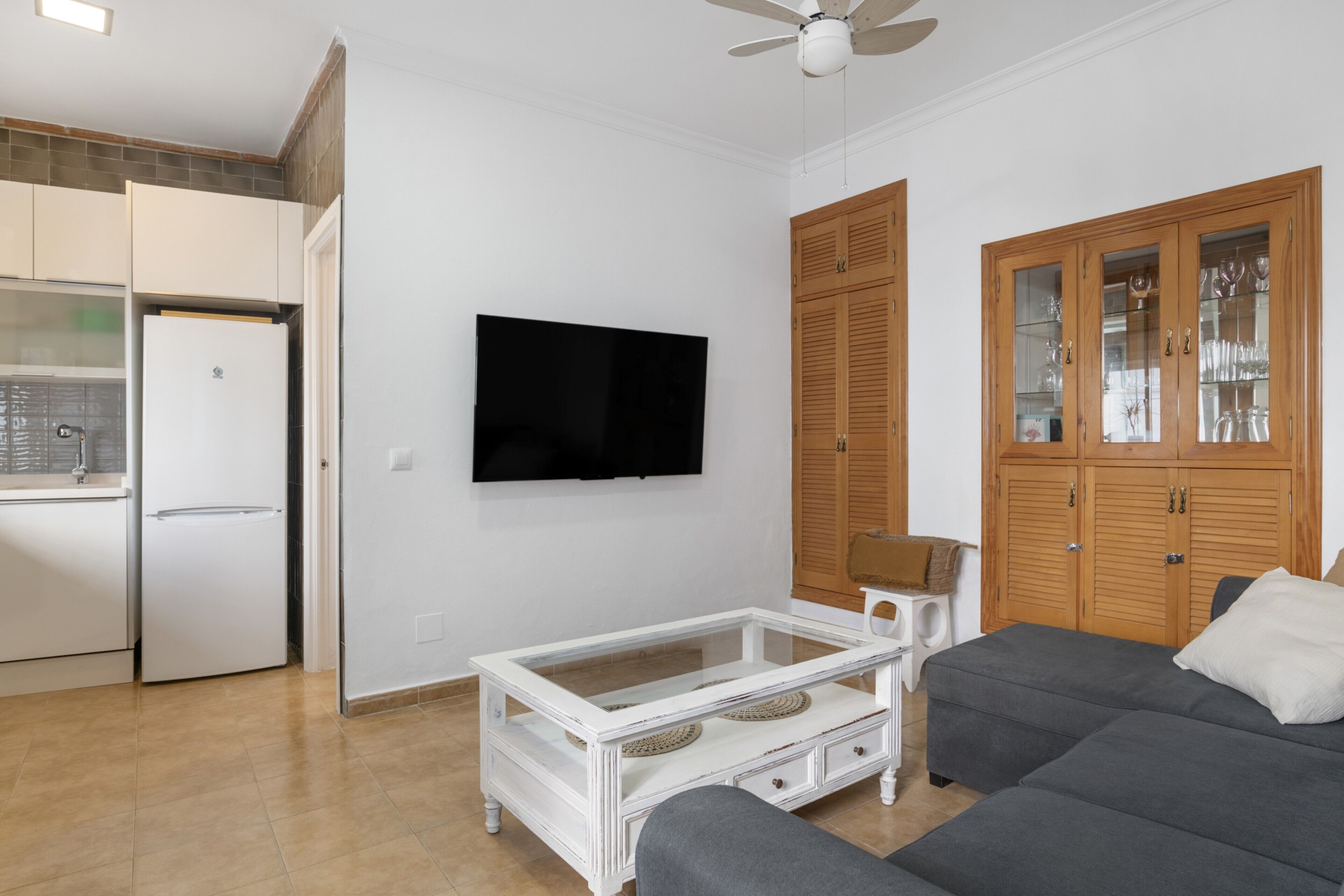 Een woonkamer met een flatscreentelevisie, een salontafel met glazen blad, een bank, houten kasten en een plafondventilator.