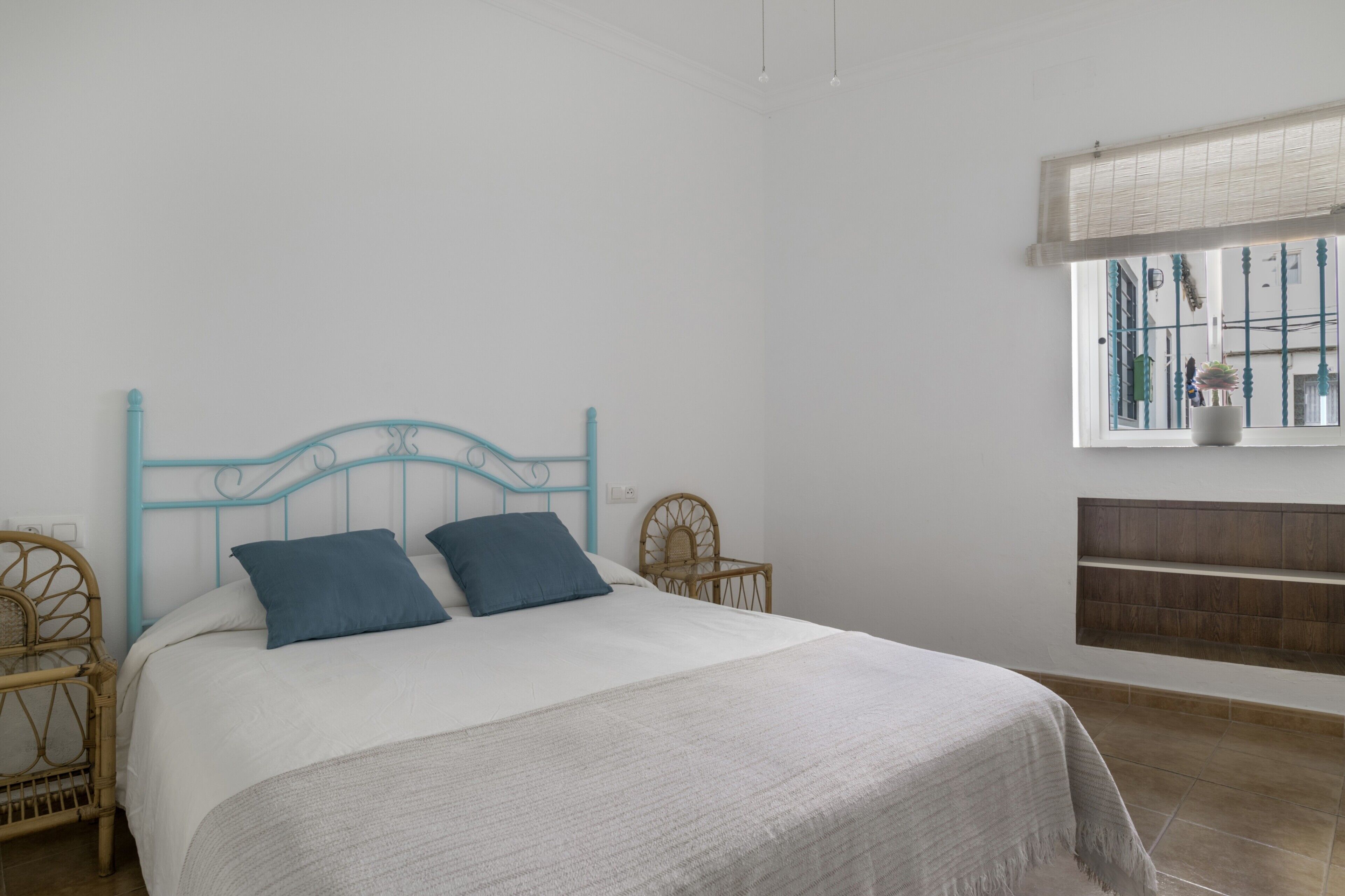 Un dormitorio con cama, dos almohadas, cabecera, ventana y estante.