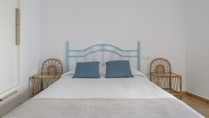 Een bed met een witte matras en twee blauwe kussens, met aan weerszijden rieten stoelen, in een kamer met witte wanden en een eenvoudig metalen hoofdbord.