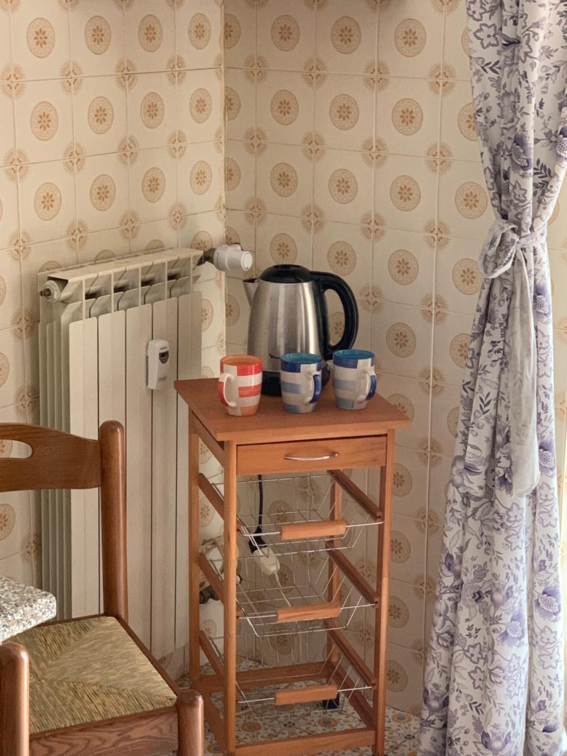 A corner of a room with a radiator, a kettle, a wooden chair, and a small wooden table with mugs and a kettle.