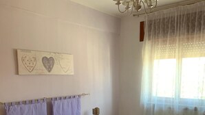 A bedroom with a bed, a nightstand, a chandelier, a window with curtains, and a wall decoration. - Apartment 'Serravalle Shopping' with Balcony and Wi-Fi (Serravalle Scrivia)