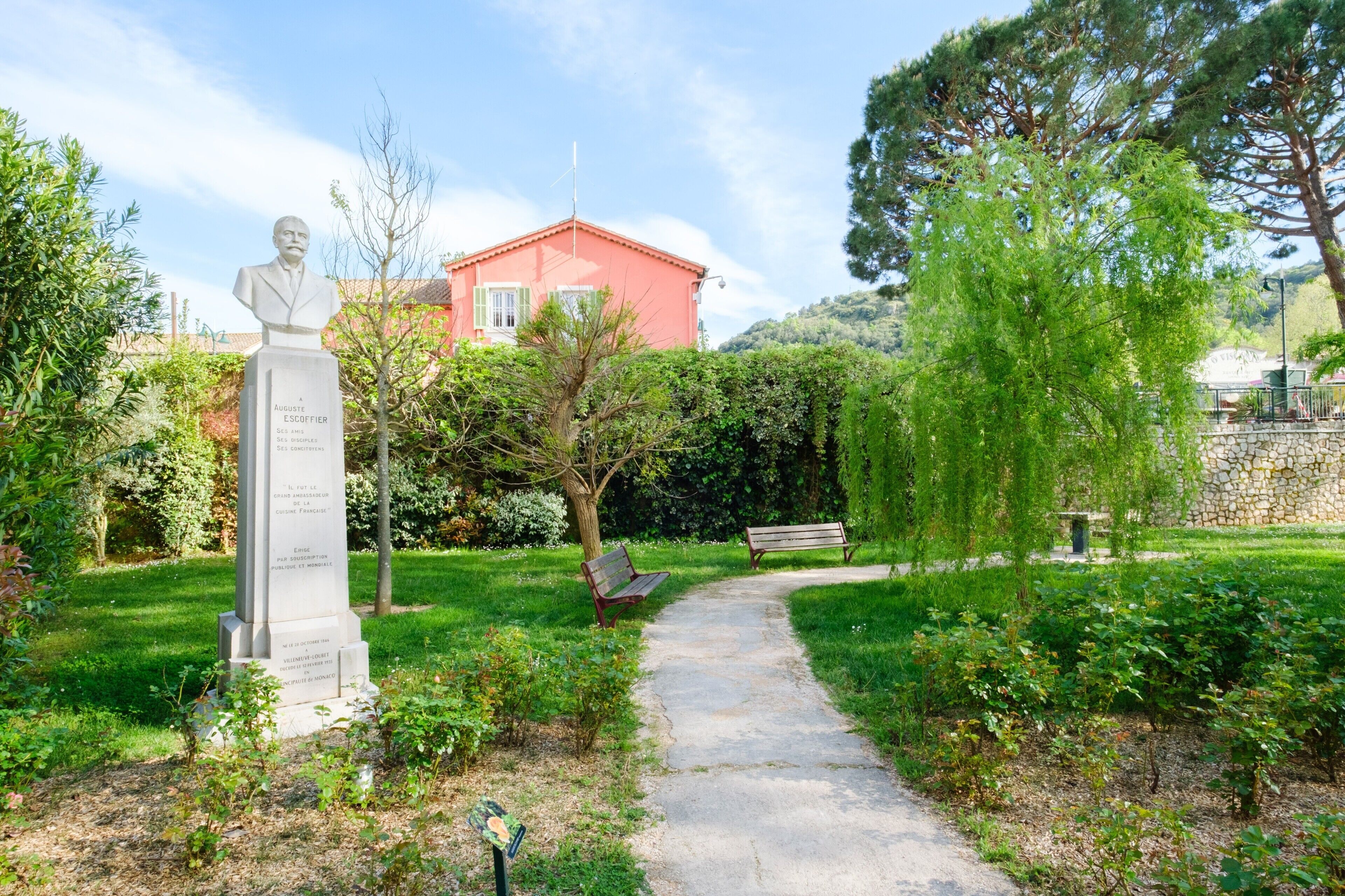 A statue in a garden with a path, benches, and a building in the background.