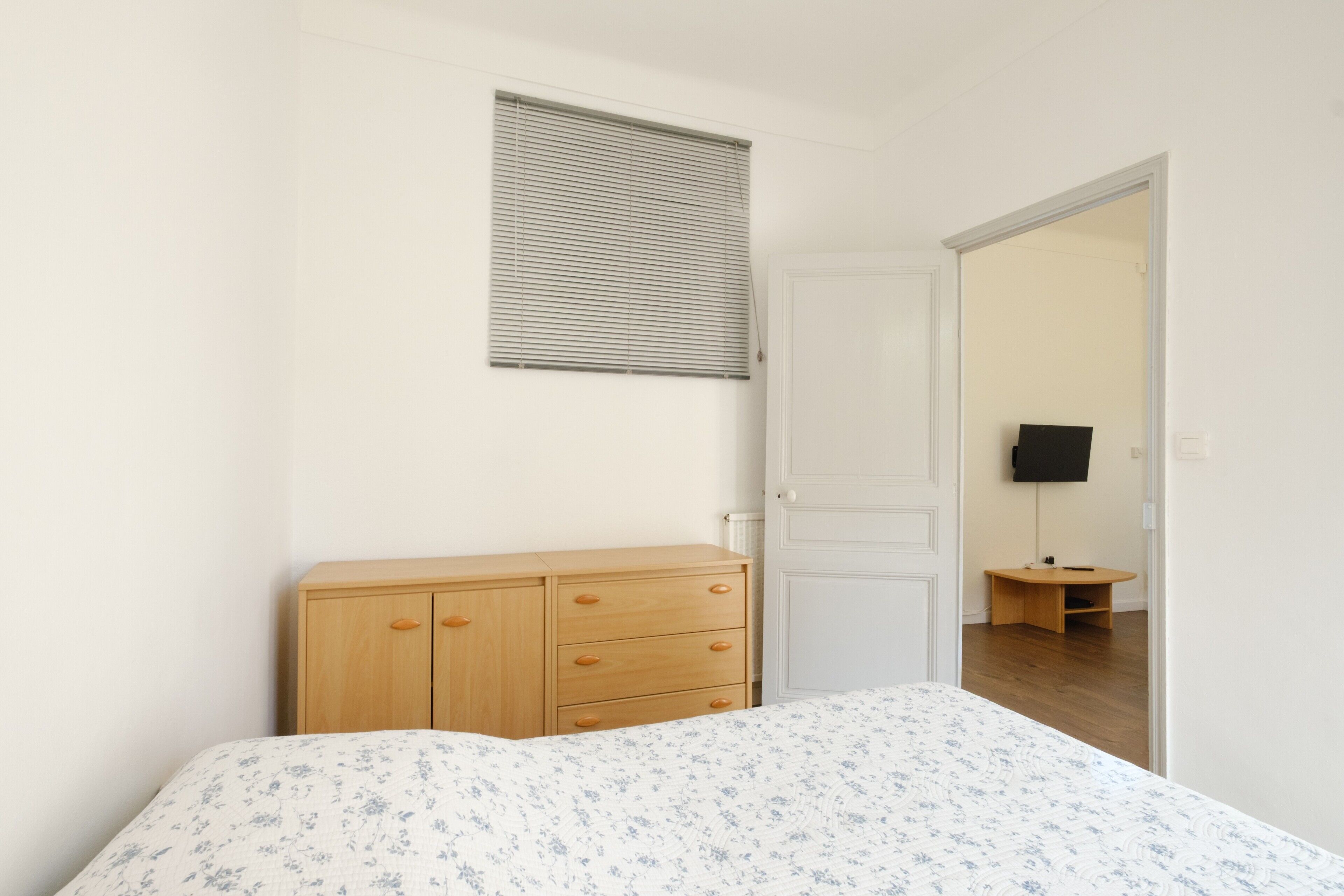 Un dormitorio con cama, cómoda de madera, puerta blanca y una pequeña mesa con televisión al fondo.