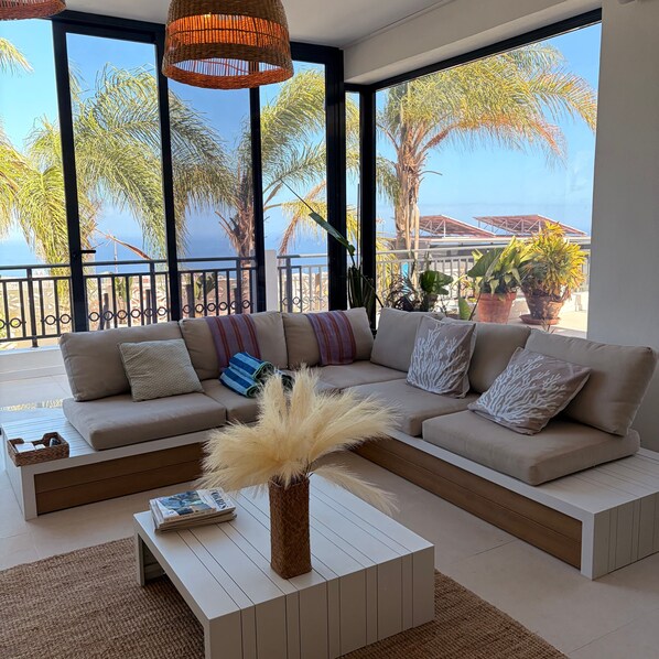 A modern living room with a large sofa, a coffee table, and a view of palm trees and the sea.