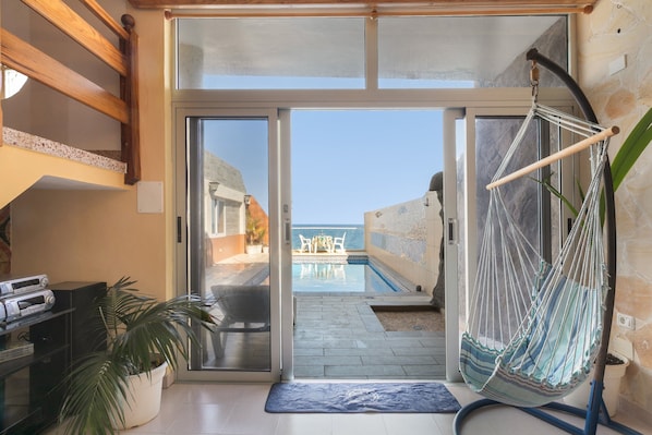 A room with a sliding glass door leading to an outdoor pool area, a hammock chair, a potted plant, and a television set on a stand.