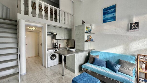 Een ruimtelijk ingerichte leefruimte met kitchenette, een wasmachine, een eethoek en een bank.