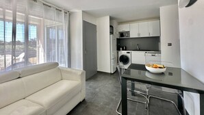 Eine moderne Wohnung mit Wohnbereich, ausgestattet mit einem weißen Sofa, einem Glastisch mit Obstschale, einer Waschmaschine und einer kompakten Küche mit weißen Schränken und einer Mikrowelle.