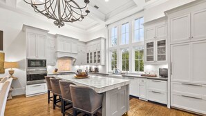 A modern kitchen with white cabinetry, a central island with bar stools, and a large window providing natural light. - Across from Sand Hill Circle Frog Pool - Golf Cart -5BR/5.5BTH Watercolor Home (Santa Rosa Beach)