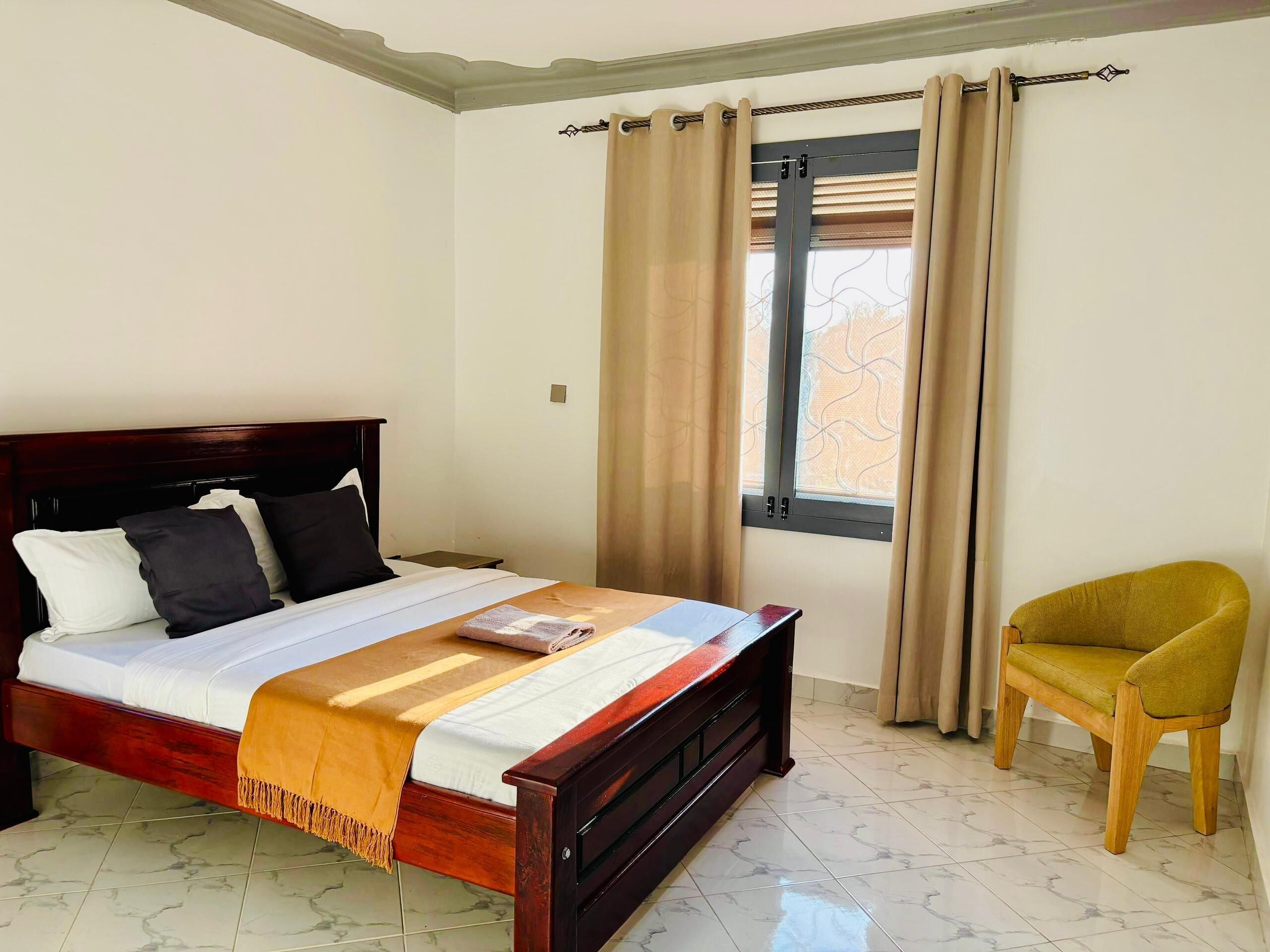 A bedroom with a wooden bed, a chair, a window with curtains, and a view of trees outside.