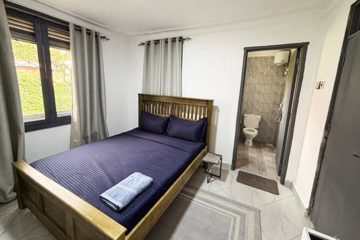 A bedroom with a wooden bed, a small bedside table, a window with curtains, and a view of the outside.