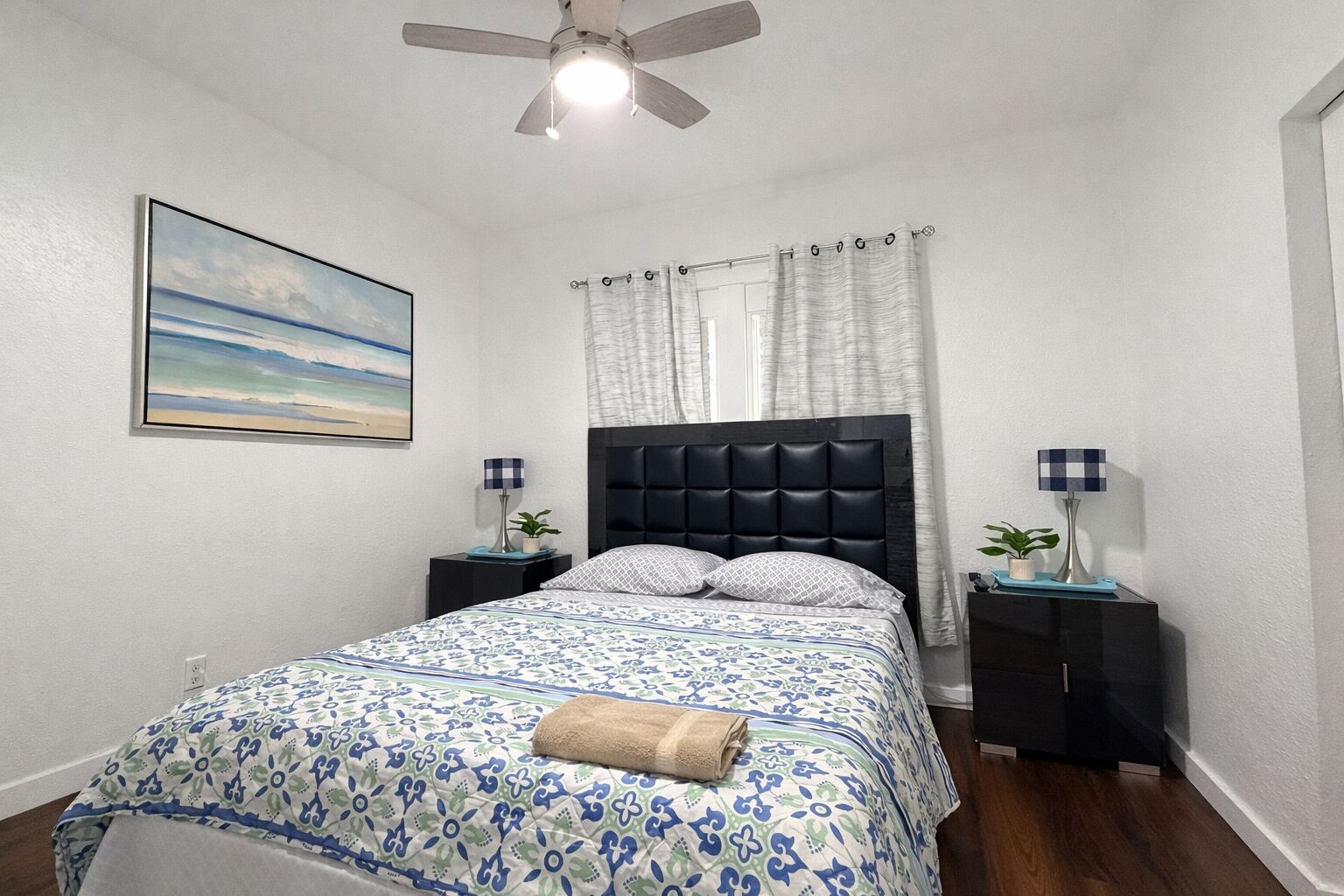 A bedroom with a bed, two bedside tables, a ceiling fan, a painting, and a window with curtains.